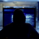 Back of hacker or cyber criminal stealing information online sitting at a computer at night. Hooded mysterious person coding spyware, malware and viruses to commit fraud, theft and phishing scam