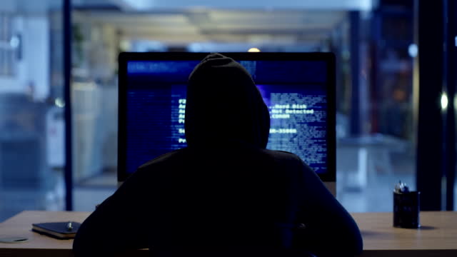 Back of hacker or cyber criminal stealing information online sitting at a computer at night. Hooded mysterious person coding spyware, malware and viruses to commit fraud, theft and phishing scam